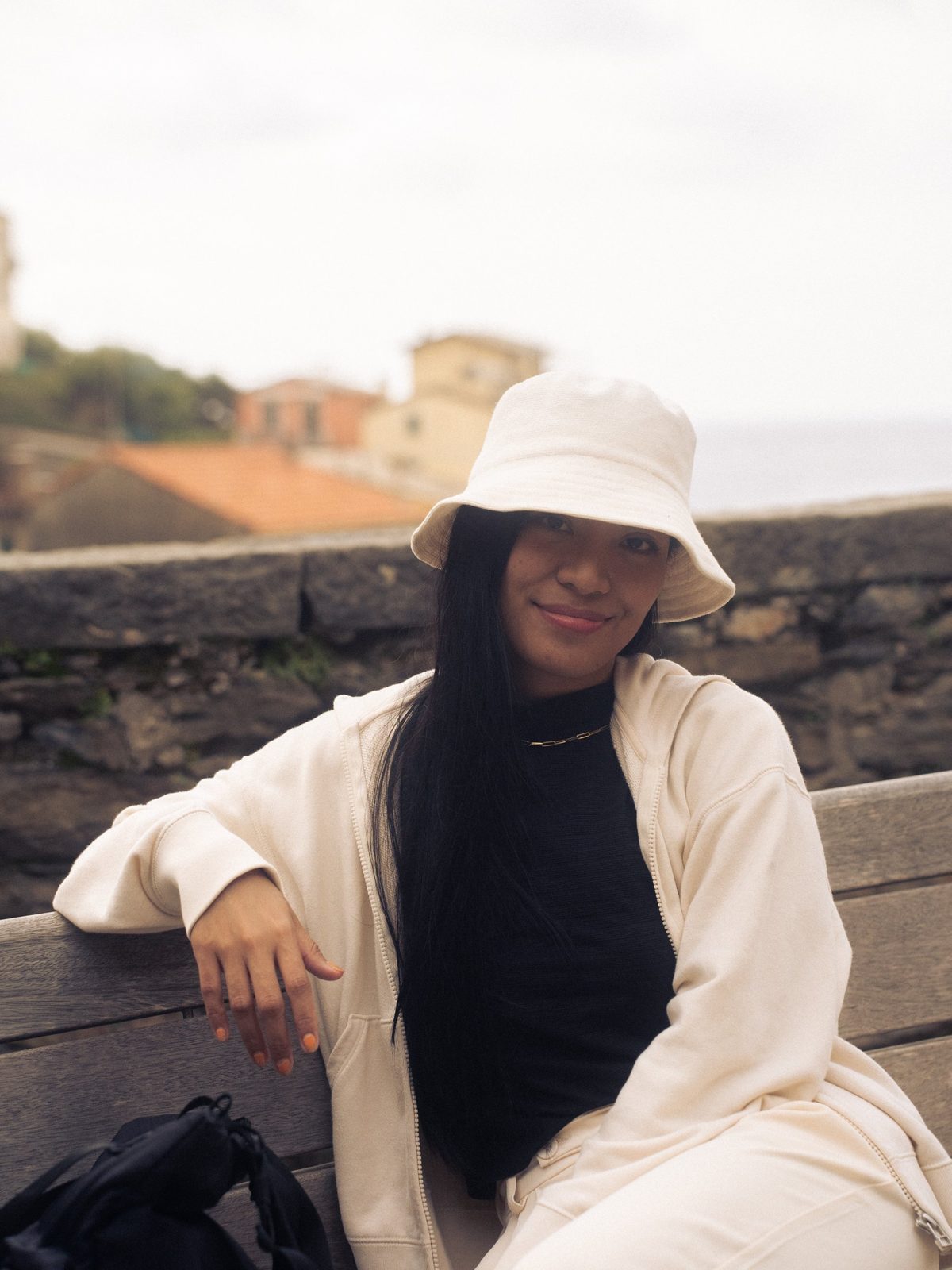 Hazel Pan in a cream bucket hat and jacket, sitting on a bench by a stone wall above a seaside town